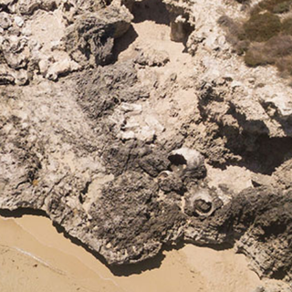Aerial view of a small sandy beach with clear water, rocky cliffs, and a striped umbrella set up on the sand.