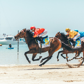 Horses racing along the shoreline during the Rockingham Beach Cup with boats anchored offshore.