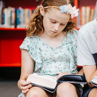 Two children sitting together in a library reading books.