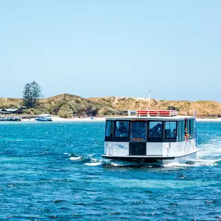 Passenger ferry travelling across clear blue water with sandy dunes and shoreline in the background.