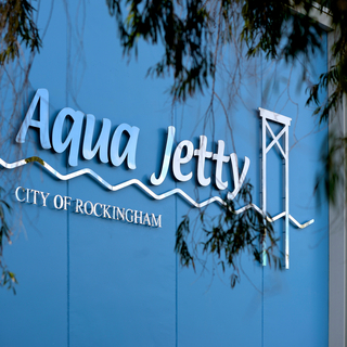 Aqua Jetty sign on a blue building wall reading ‘Aqua Jetty – City of Rockingham,’ partially framed by tree branches.