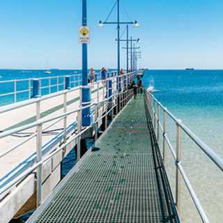 View from a jetty overlooking people enjoying the shallow waters at the beach with moored boats in the background.