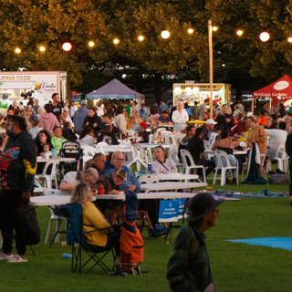 Large outdoor community event with people gathered around tables, food stalls and festoon lights at dusk.