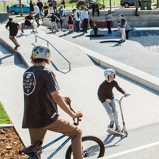 Young people riding scooters and BMX bikes at a busy skate park with ramps and rails.