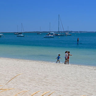 Sunny beach with boats anchored offshore and people walking along the sand.