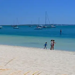 Sunny beach with boats anchored offshore and people walking along the sand.