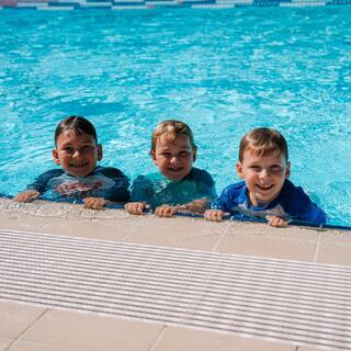 Three children holding onto the edge of an outdoor swimming pool during a swim session.