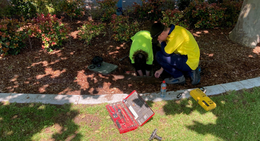 Two youth trainees working together on a maintenance task in a garden bed with tools laid out nearby.