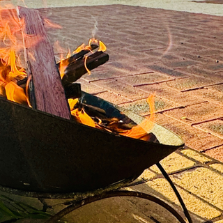 A smoking ceremony fire burning in a metal bowl with green leaves beside it on paved ground
